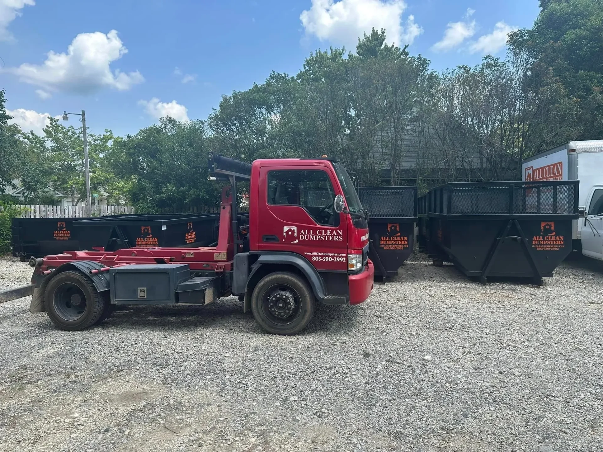 A red truck parked in the gravel next to a dumpster.